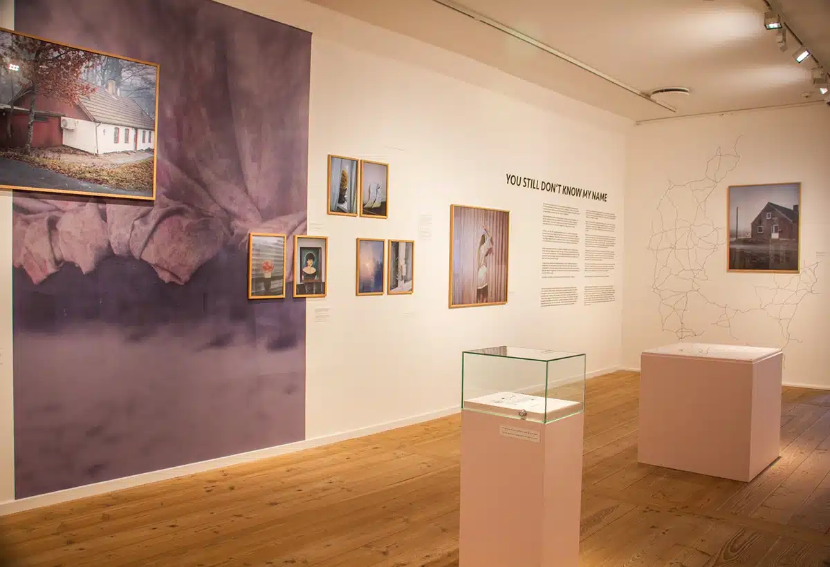 Et lyst gallerirum i udstillingen ”You still don’t know my name” på KØN med titlen skrevet på væggen, en glasmontre i forgrunden og et værk med et kort over landeveje i baggrunden