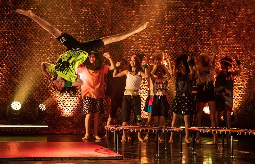 Dreng laver salto over trampolinen til Flying Superkids show, mens en flok børn i farverigt hverdagstøj danser og hepper Dreng laver salto over trampolinen til Flying Superkids show, mens en flok børn i farverigt hverdagstøj danser og hepper