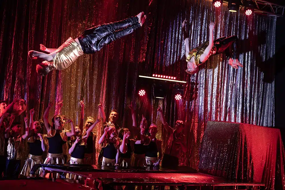 To Flying Superkids gymnaster flyver vandret gennem luften i guldkostumer, mens holdet hepper på dem i rødt scenelys To Flying Superkids gymnaster flyver vandret gennem luften i guldkostumer, mens holdet hepper på dem i rødt scenelys